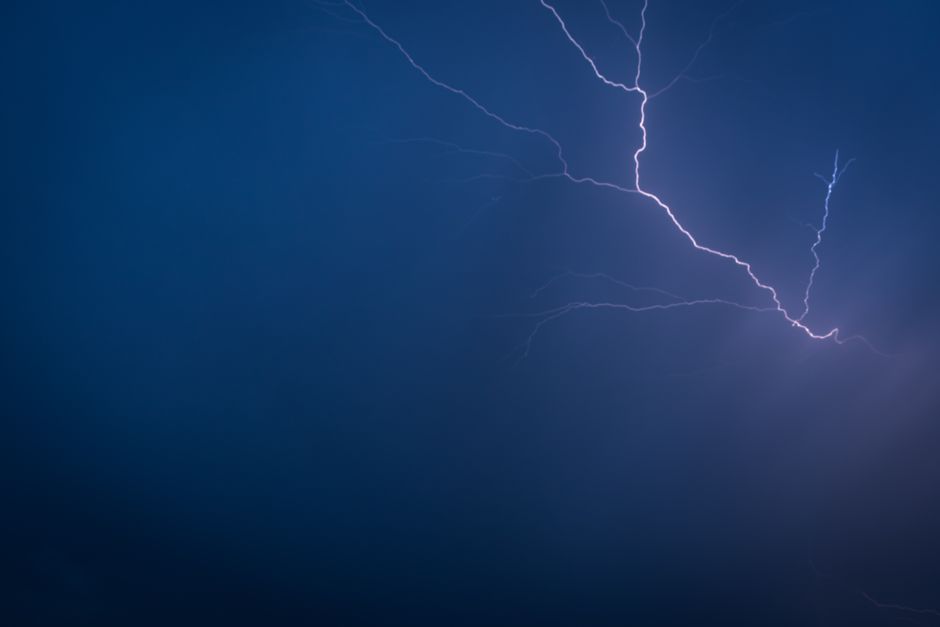 The image shows a dark night sky illuminated by a vivid lightning strike, with jagged, branching bolts piercing through the cloudless darkness. The lightning appears bright white with subtle bluish ti
